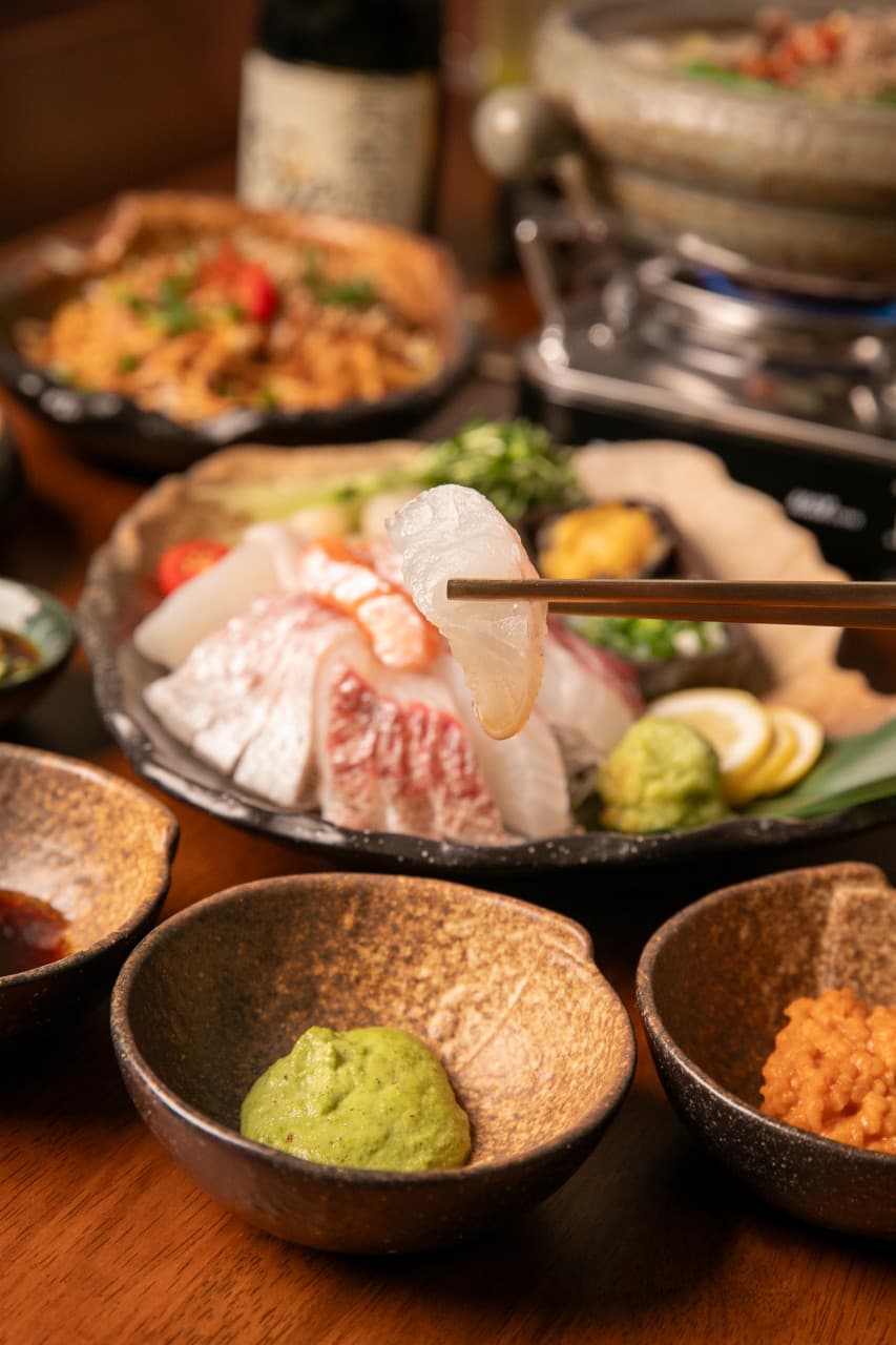 나무 테이블 위 젓가락으로 집어 올린 회 한 점과 사시미 플레이트, 와사비 종지 및 1인용 전골 차림