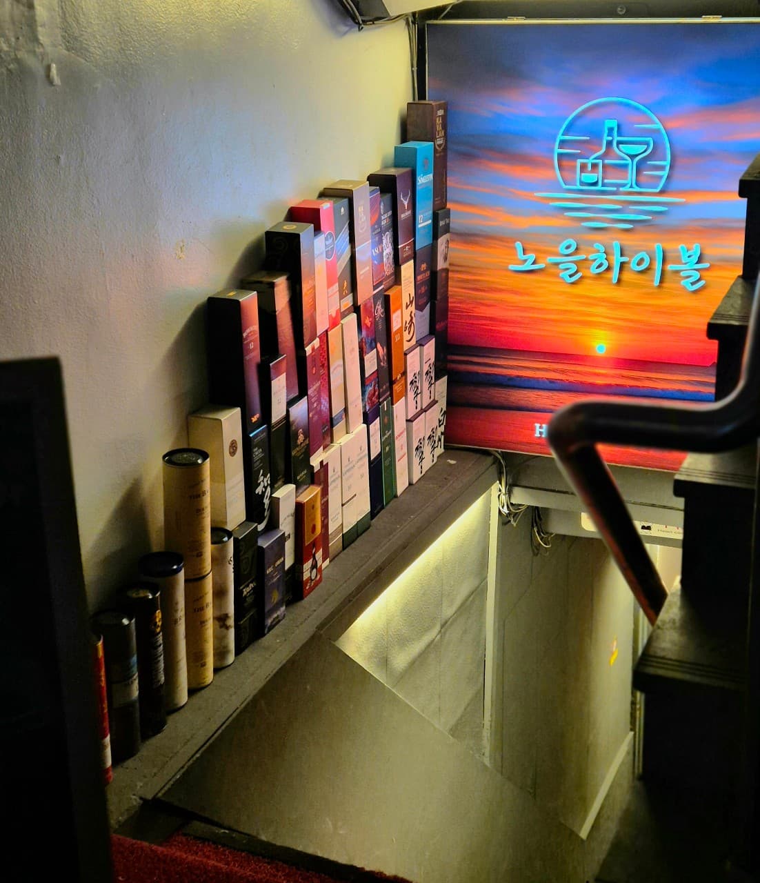 지하로 이어지는 좁은 계단 옆벽에 일렬로 진열된 주류 상자들과 석양 풍경의 조명 간판