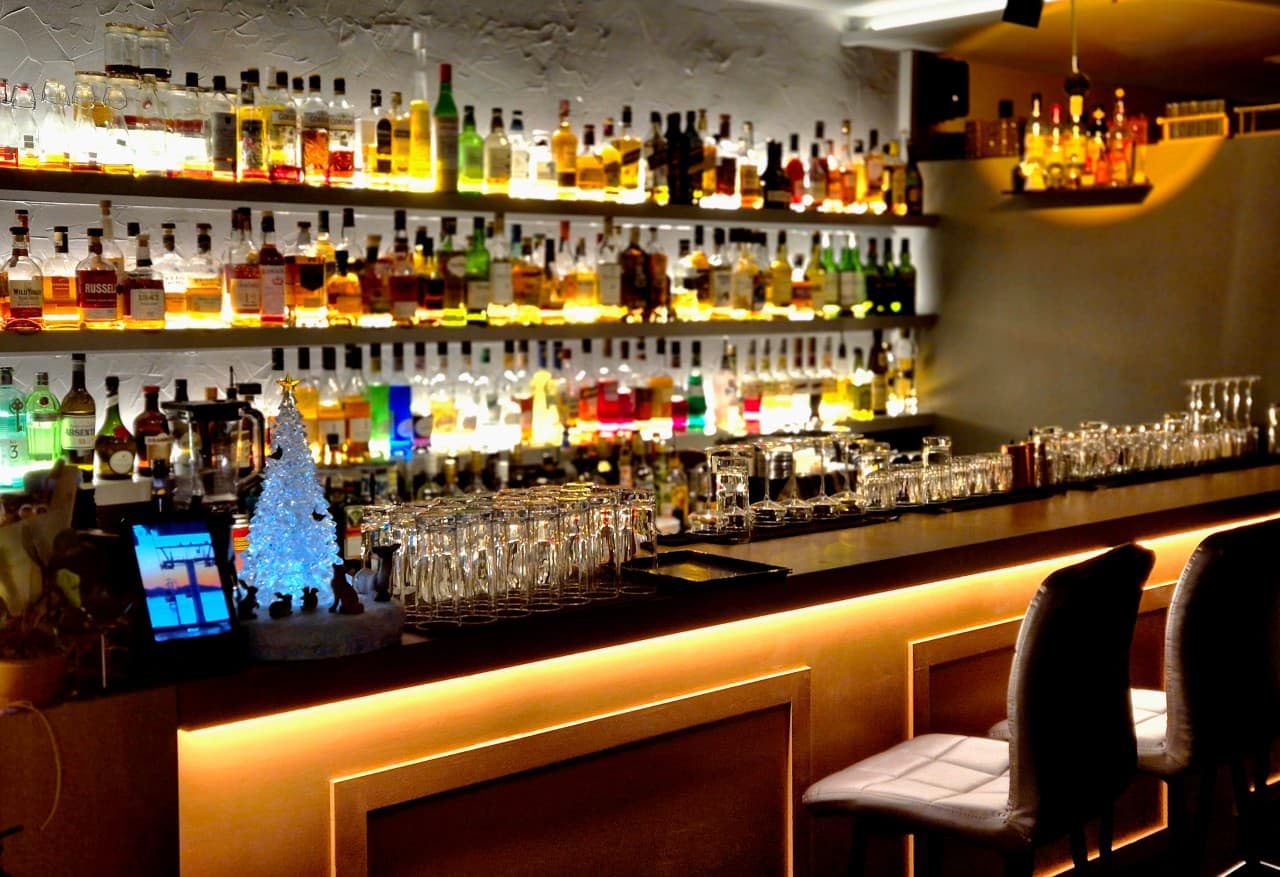 노란 조명이 켜진 바 카운터 테이블과 일렬로 배치된 1인석 의자 및 술병 선반