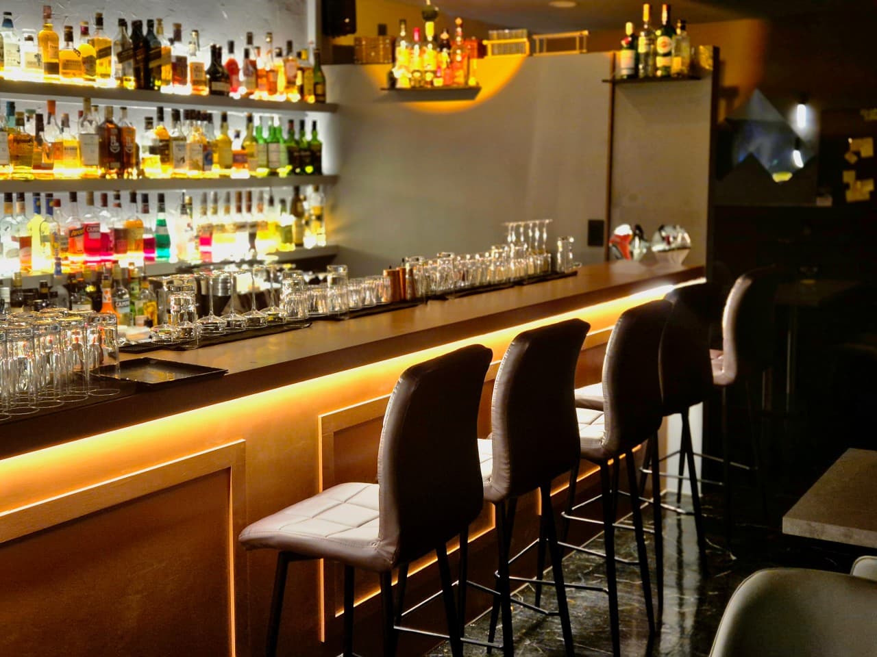노란 간접 조명이 들어오는 긴 카운터 테이블과 일렬로 배치된 1인용 바 의자, 벽면의 다양한 술병 선반