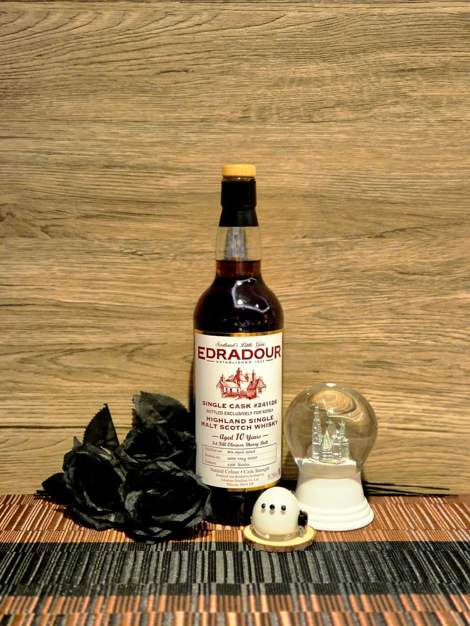 나무 질감의 벽면 앞 위스키 병과 검은 장미, 스노우볼이 나란히 놓인 바 테이블의 1인석 전경