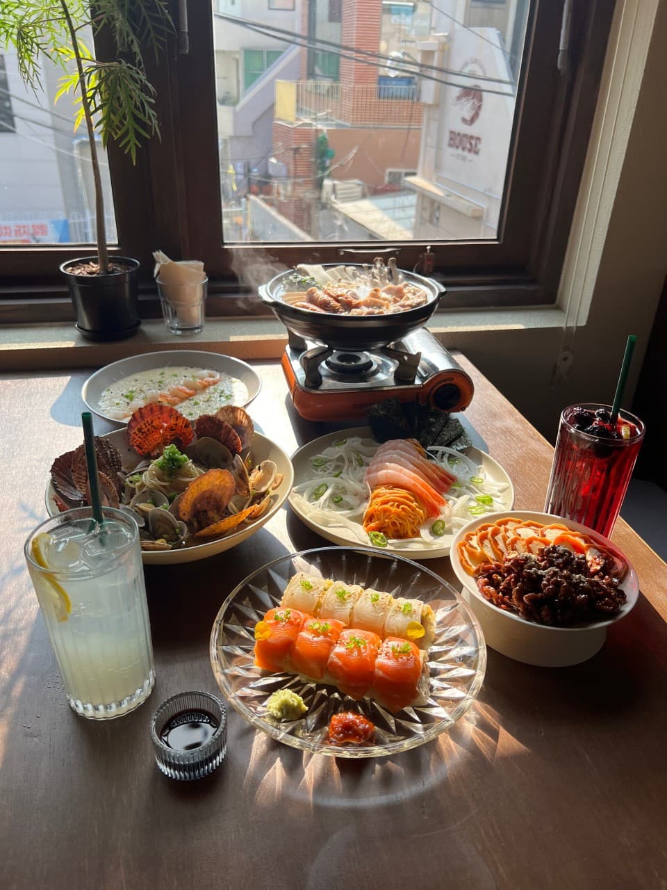 창가 나무 테이블 위 김이 나는 나베와 연어 롤, 해산물 파스타와 하이볼 잔이 놓인 모습