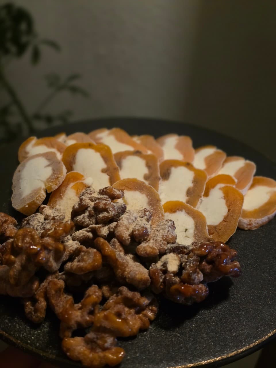 낮은 조도의 실내, 검은 원형 접시에 담긴 곶감 크림치즈 말이와 설탕 가루를 묻힌 호두 강정