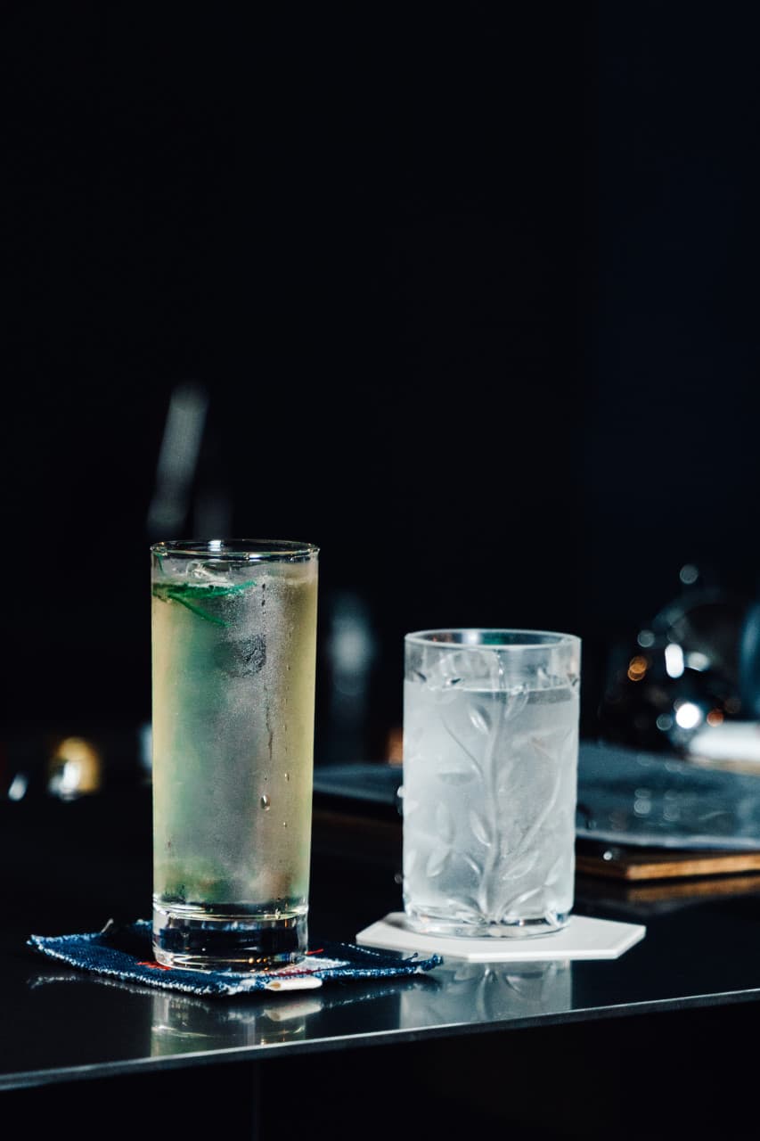 어두운 실내 검은색 바 카운터 위 데님 코스터에 놓인 하이볼 잔과 얼음이 담긴 나뭇잎 무늬 유리잔