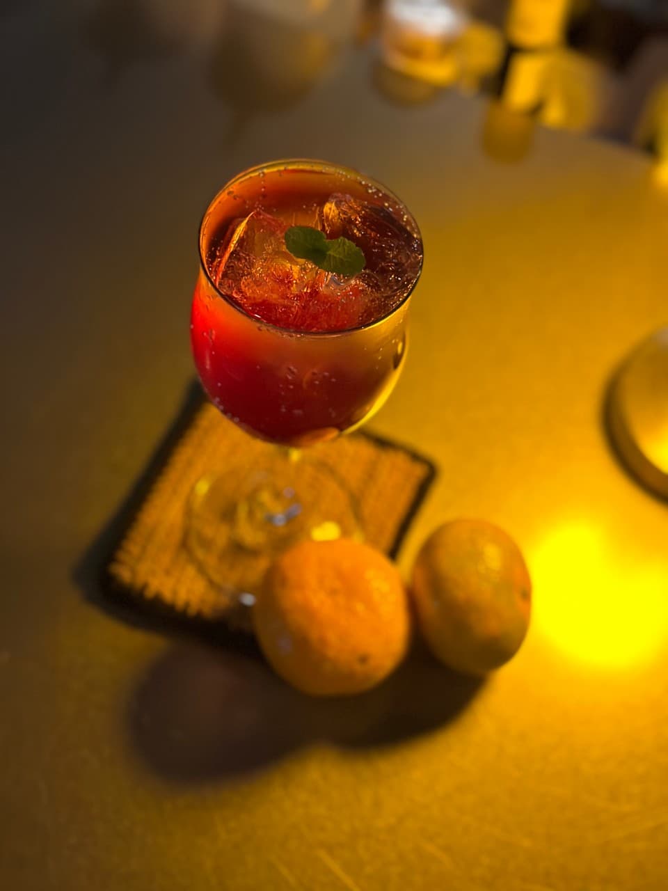 노란 조명이 비치는 어두운 바 테이블 위 코스터에 놓인 붉은 색 칵테일 한 잔과 귤 두 알