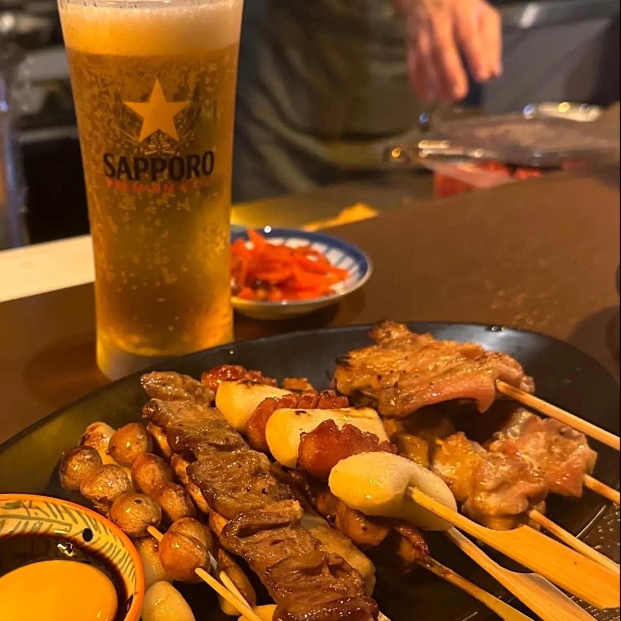 나무 카운터 위 검은 접시에 담긴 닭꼬치와 은행구이, 계란 노른자 소스 그리고 생맥주 잔이 놓인 1인용 좌석 전경