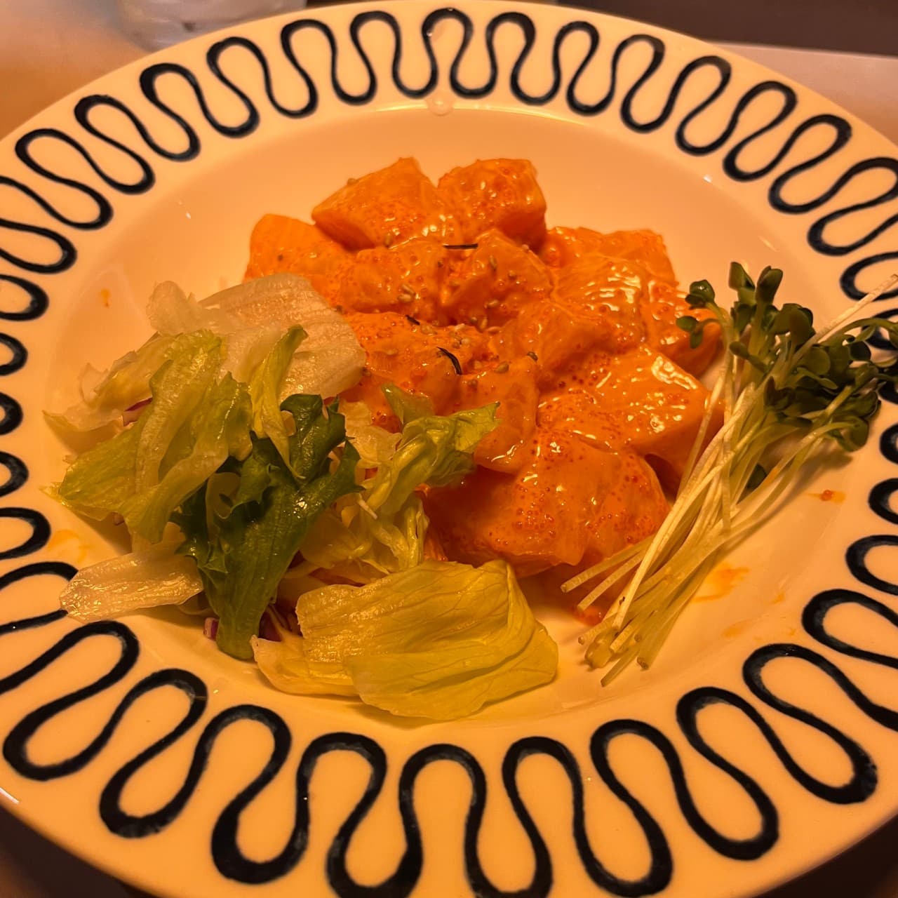 주황색 조명 아래 물결무늬 접시에 담긴 명란 소스 연어 버무림과 양상추 샐러드, 무순이 곁들여진 1인용 안주