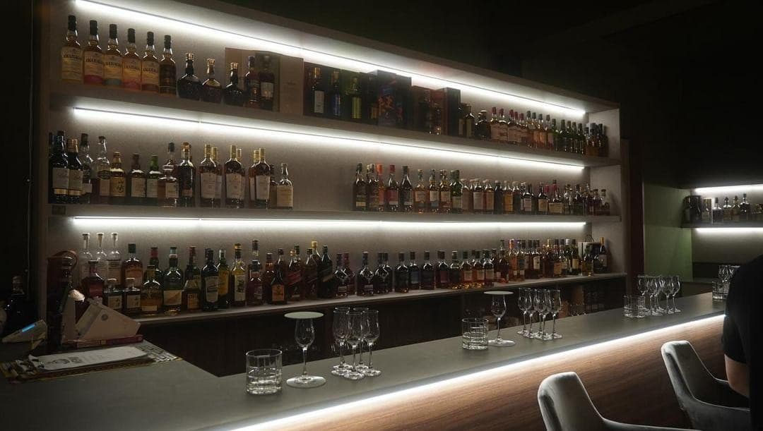 수평 조명이 비추는 술병 선반 앞 여러 종류의 글라스가 놓인 바 카운터와 1인용 좌석