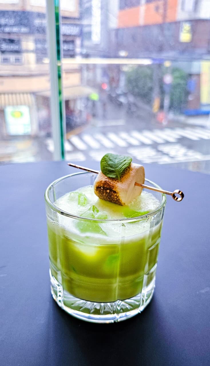횡단보도가 내려다보이는 창가 좌석 위 구운 마시멜로와 민트 잎이 얹어진 연두색 칵테일과 유리창 밖 거리 전경