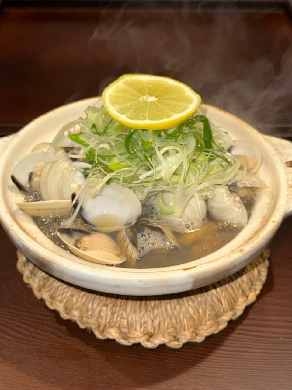 김이 올라오는 조개탕 뚝배기 위에 가득 쌓인 파채와 레몬 슬라이스, 짚으로 짠 냄비 받침대와 나무 카운터 테이블