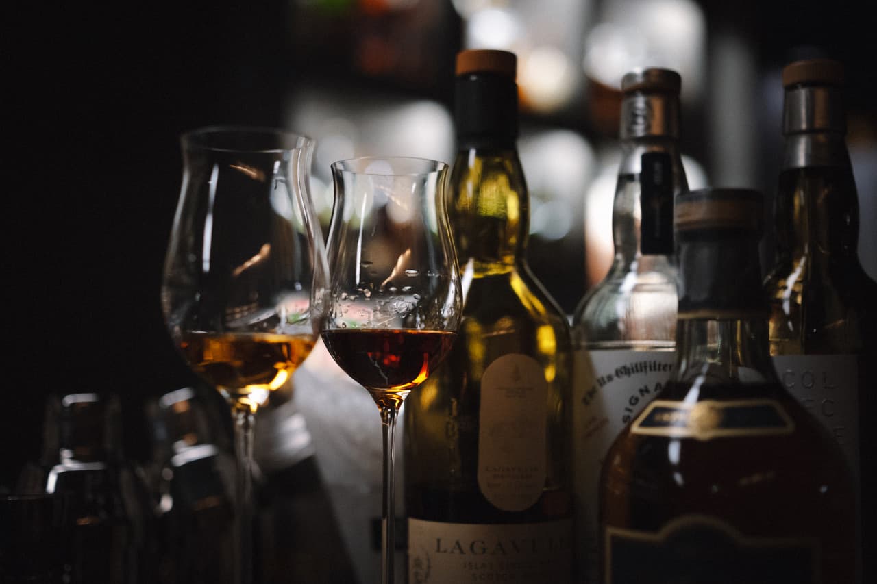 어두운 조명 아래 호박색 술이 담긴 두 잔의 테이스팅 글라스와 뒤로 줄지어 선 위스키 병들