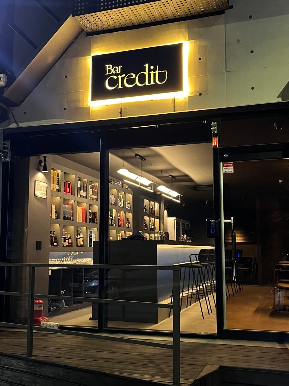 노란 백라이트 간판 아래 통유리 너머로 보이는 긴 검은색 카운터 테이블과 높은 의자, 술병들이 가득한 벽면 진열장