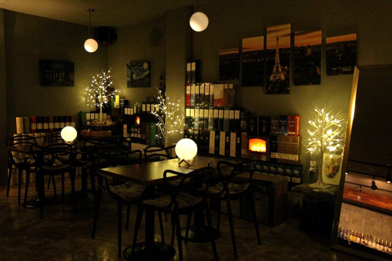 어두운 조명 아래 놓인 소규모 테이블과 의자, 벽면의 주류 박스들과 구형 램프 및 트리 형태의 전구