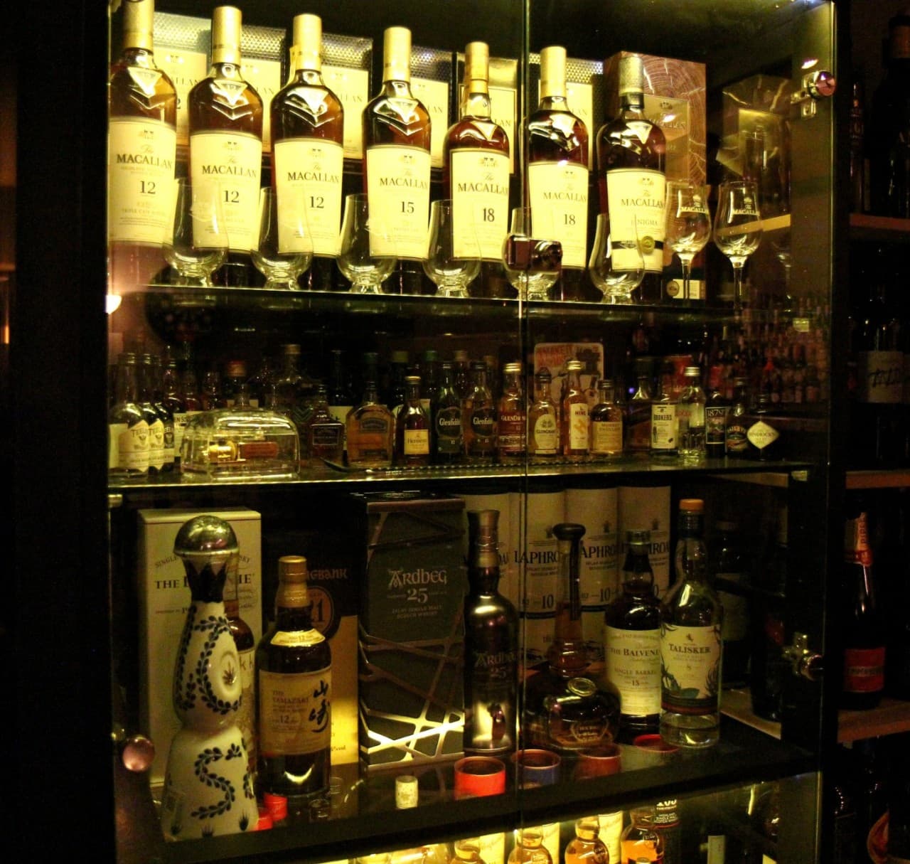 어두운 조명이 비추는 유리 장식장 내부에 진열된 맥캘란과 야마자키 등 수십 종류의 위스키 병과 글렌캐런 잔