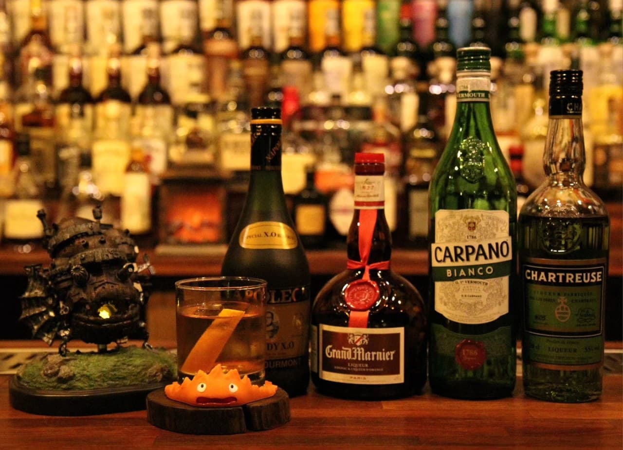어두운 조명 아래 목재 바 테이블 위 칵테일 잔과 캐릭터 소품, 배경에 가득 찬 다양한 리큐르 병들이 보이는 전경