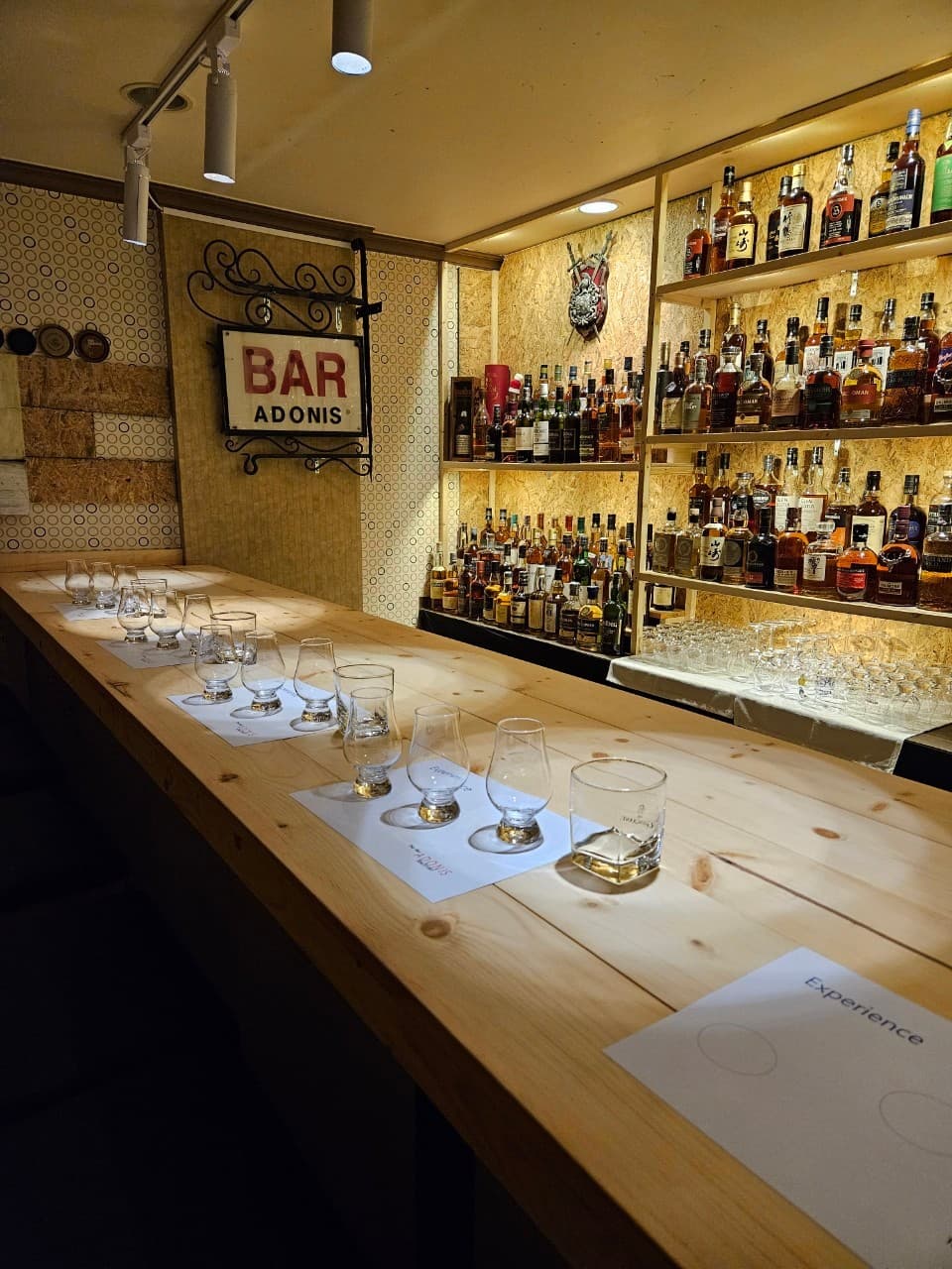 밝은 색 나무 카운터 위 일렬로 놓인 위스키 잔들과 술병이 가득한 벽면 선반 전경