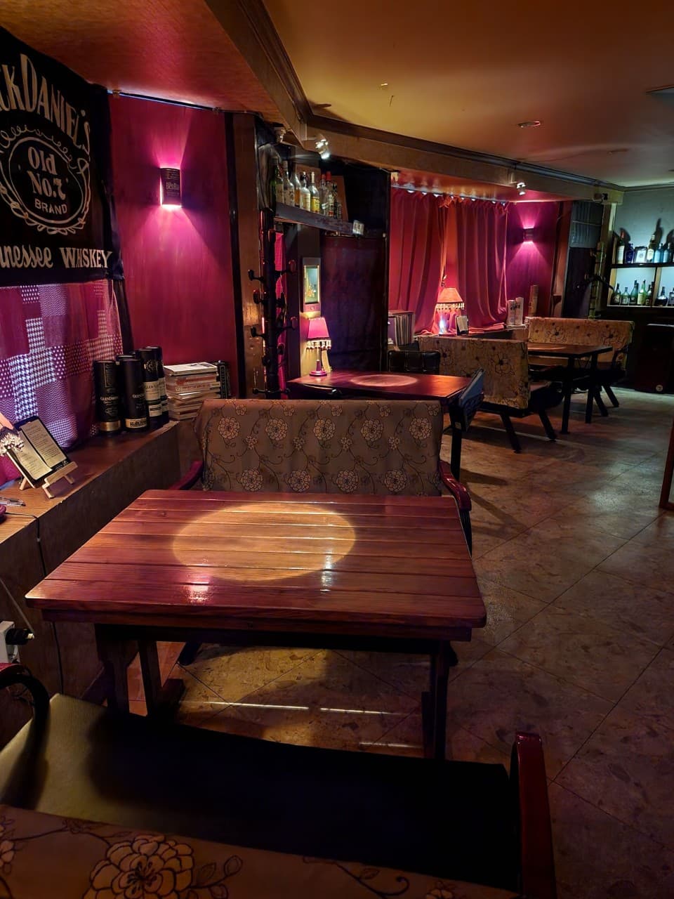 어두운 실내 붉은 조명 아래 개별 핀 조명이 켜진 목재 테이블과 패턴 소파, 벽면에 진열된 술병들