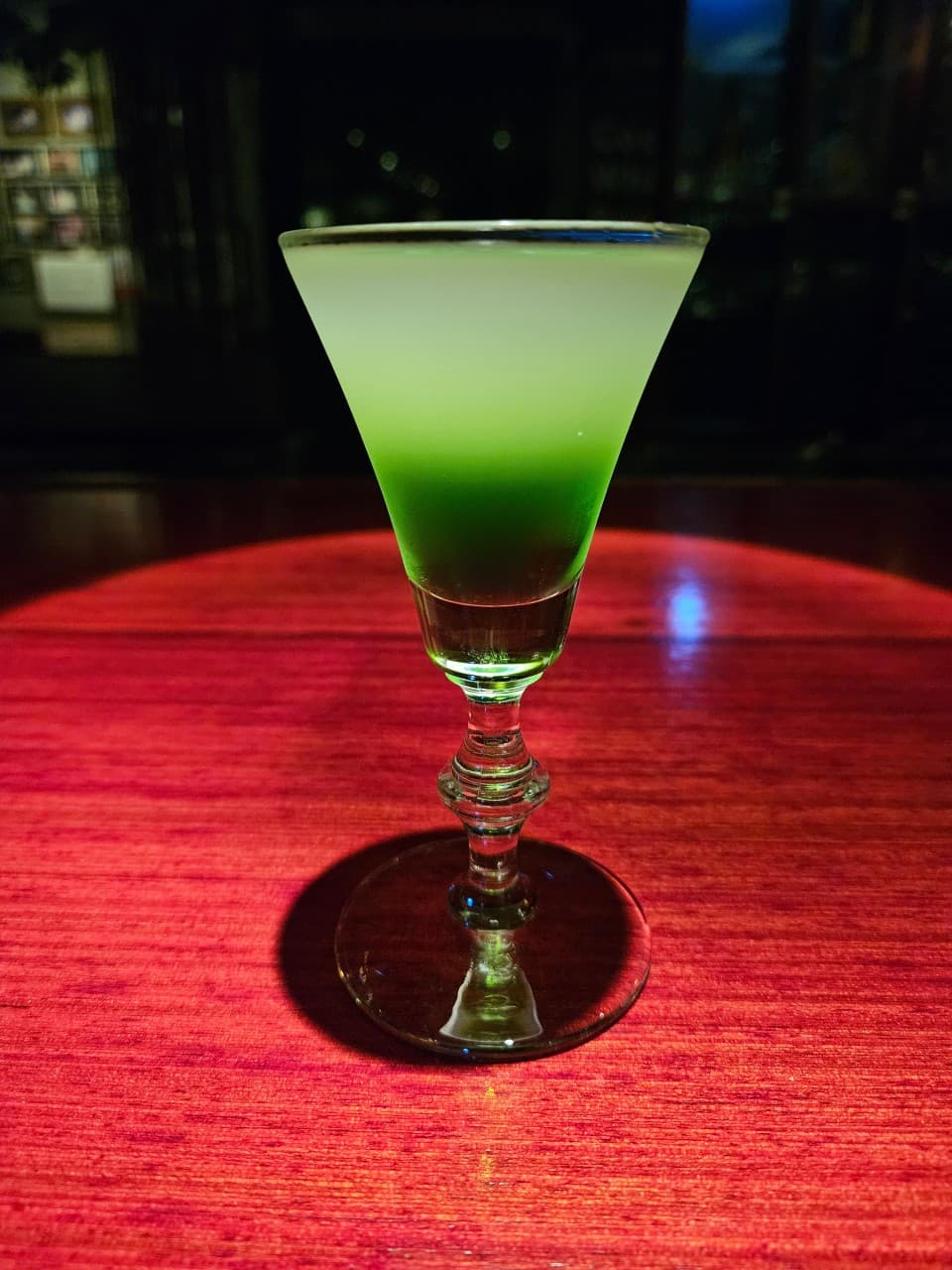 붉은 조명이 비치는 어두운 목재 바 테이블 위 두 층으로 나뉜 연두색 칵테일 한 잔
