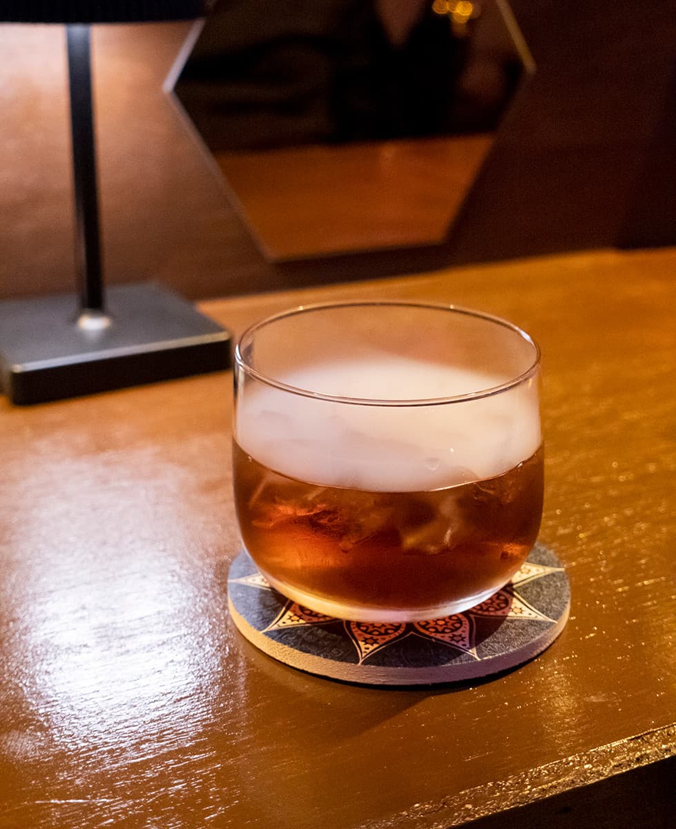 어두운 조명 아래 목재 테이블 위 흰 연기가 담긴 칵테일 잔과 기하학 문양의 코스터