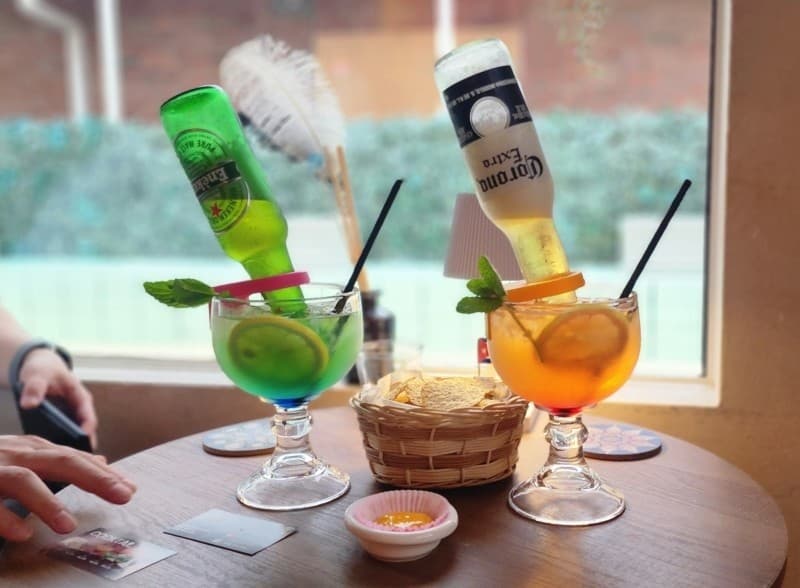 창가 나무 테이블 위 맥주병이 거꾸로 꽂힌 두 종류의 칵테일과 레몬 슬라이스, 바구니에 담긴 나초 칩