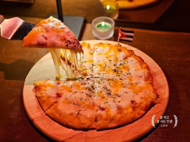 어두운 조명 아래 나무 테이블 위 원형 판에 담긴 치즈 피자와 서버로 들어 올린 한 조각, 작은 깃발 장식과 초록색 캔들