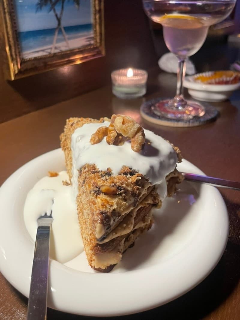 어두운 조명 아래 목재 테이블 위 크림과 견과류를 얹은 케이크, 긴 줄기의 칵테일 잔과 작은 촛불