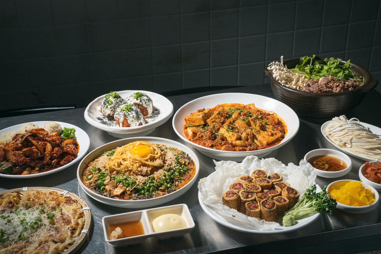 어두운 타일 배경의 금속 테이블 위 계란 노른자가 올라간 비빔면과 전골, 육회말이 등 여러 종류의 1인 안주 차림