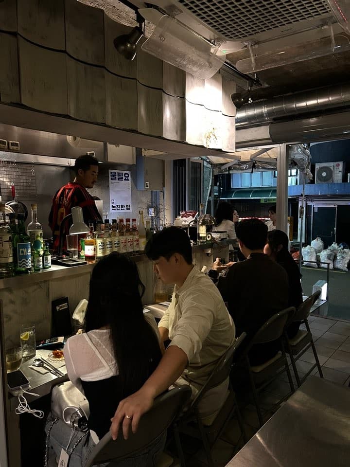 어두운 조명 아래 긴 카운터 석에 앉은 손님들과 선반 위 다양한 술병이 늘어선 주방 전경