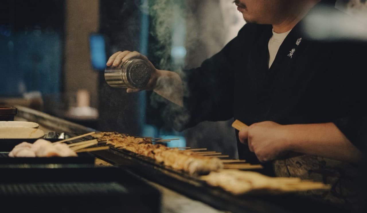 어두운 조명 아래 카운터 너머로 숯불 위 닭꼬치에 시즈닝을 뿌리는 조리사와 피어오르는 연기