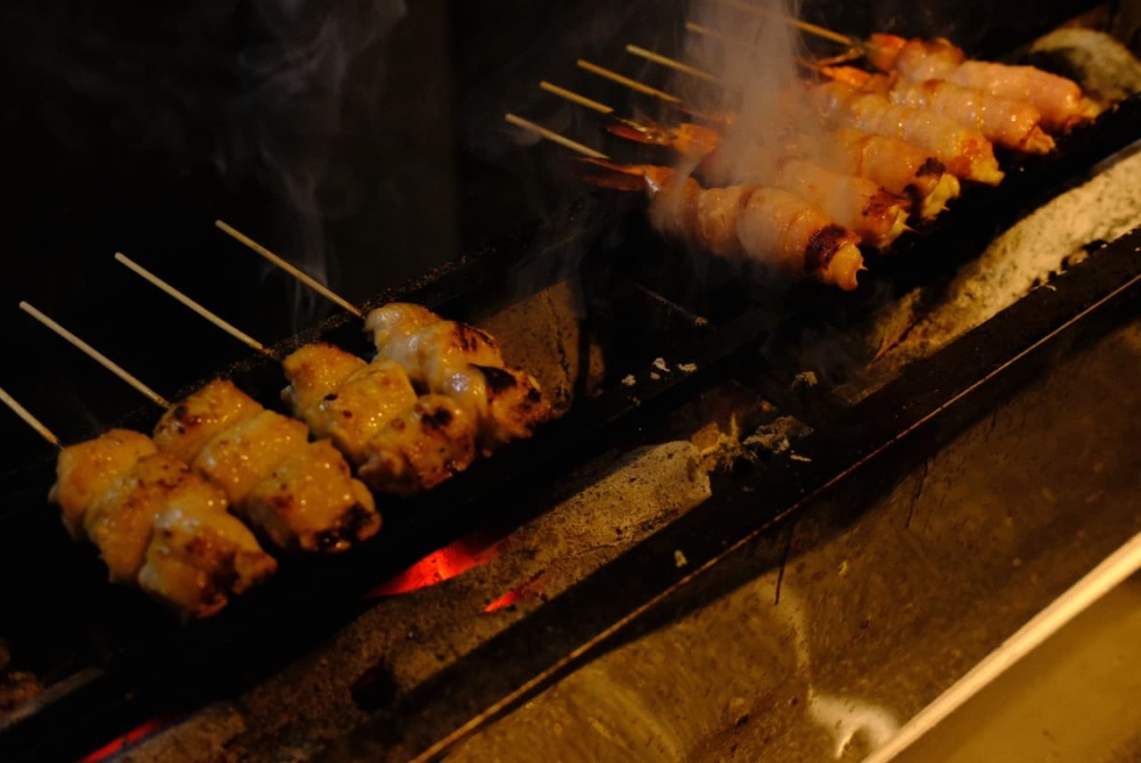 어두운 조명 아래 붉은 숯불 위에서 연기를 내며 익어가는 닭꼬치와 베이컨 말이 꼬치구이