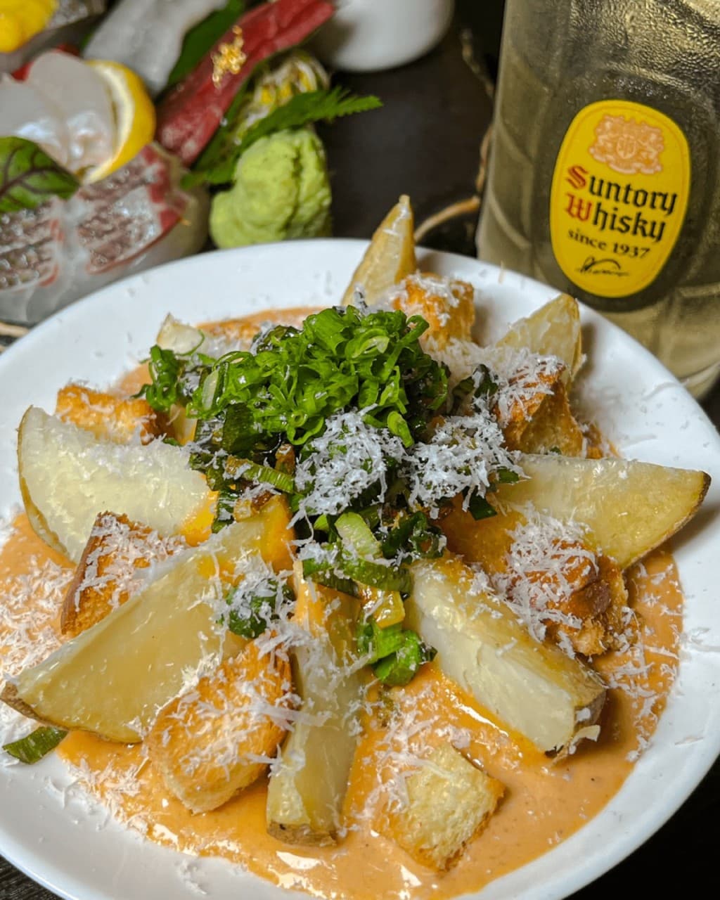 주황색 소스와 치즈 가루가 뿌려진 감자 요리 접시 옆에 놓인 산토리 위스키 하이볼 잔과 생고추냉이