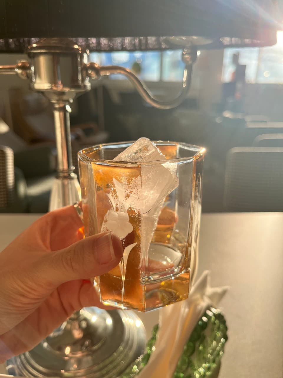 햇빛이 비치는 실내 창가 좌석에서 한 손으로 들고 있는 커다란 얼음 조각과 갈색 액체가 담긴 유리잔