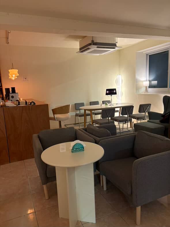 노란색 조명 아래 목재 카운터와 1인용 회색 소파가 배치된 흰색 원형 탁자 및 배경의 긴 테이블 좌석