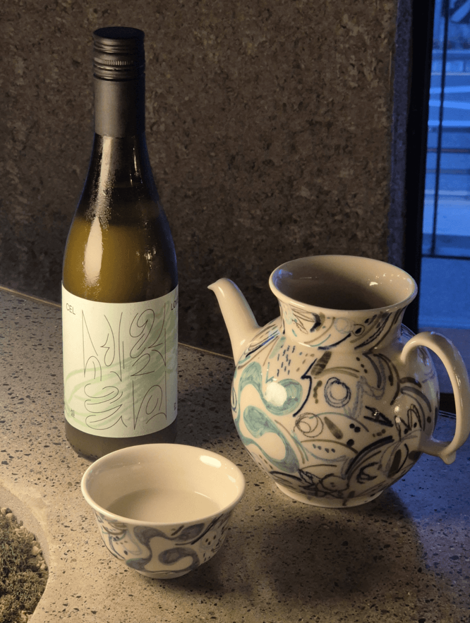 어두운 실내 조명 아래 석재 테이블 위 초록색 술병과 푸른 문양의 도자기 주전자 및 술이 담긴 잔