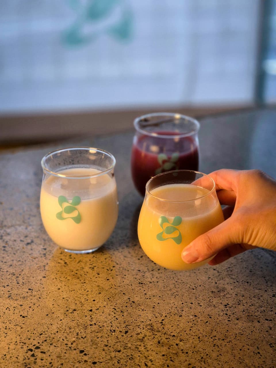 석재 바 테이블 위에 놓인 세 종류의 음료와 로고가 새겨진 잔을 쥐고 있는 1인의 손