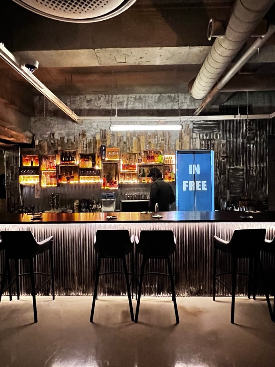 노출 배관 천장 아래 조명이 켜진 주류 선반과 바 카운터에 일렬로 배치된 1인용 의자들