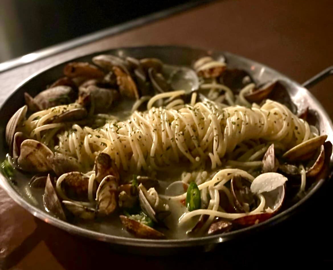 어두운 조명의 나무 카운터 위 금속 접시에 담긴 바지락 파스타와 썰어 넣은 고추 및 허브 가루