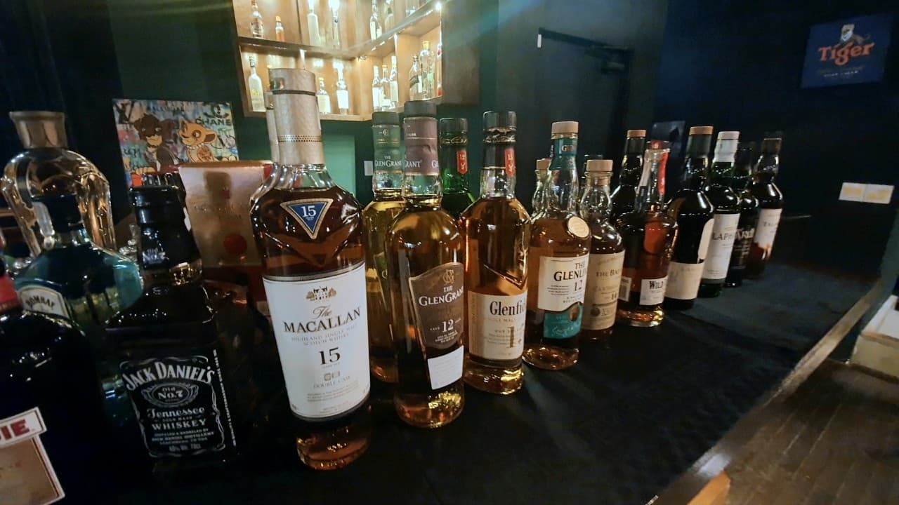 검은색 바 카운터 위에 일렬로 진열된 다양한 종류의 위스키 병들과 조명이 켜진 목재 술 진열장