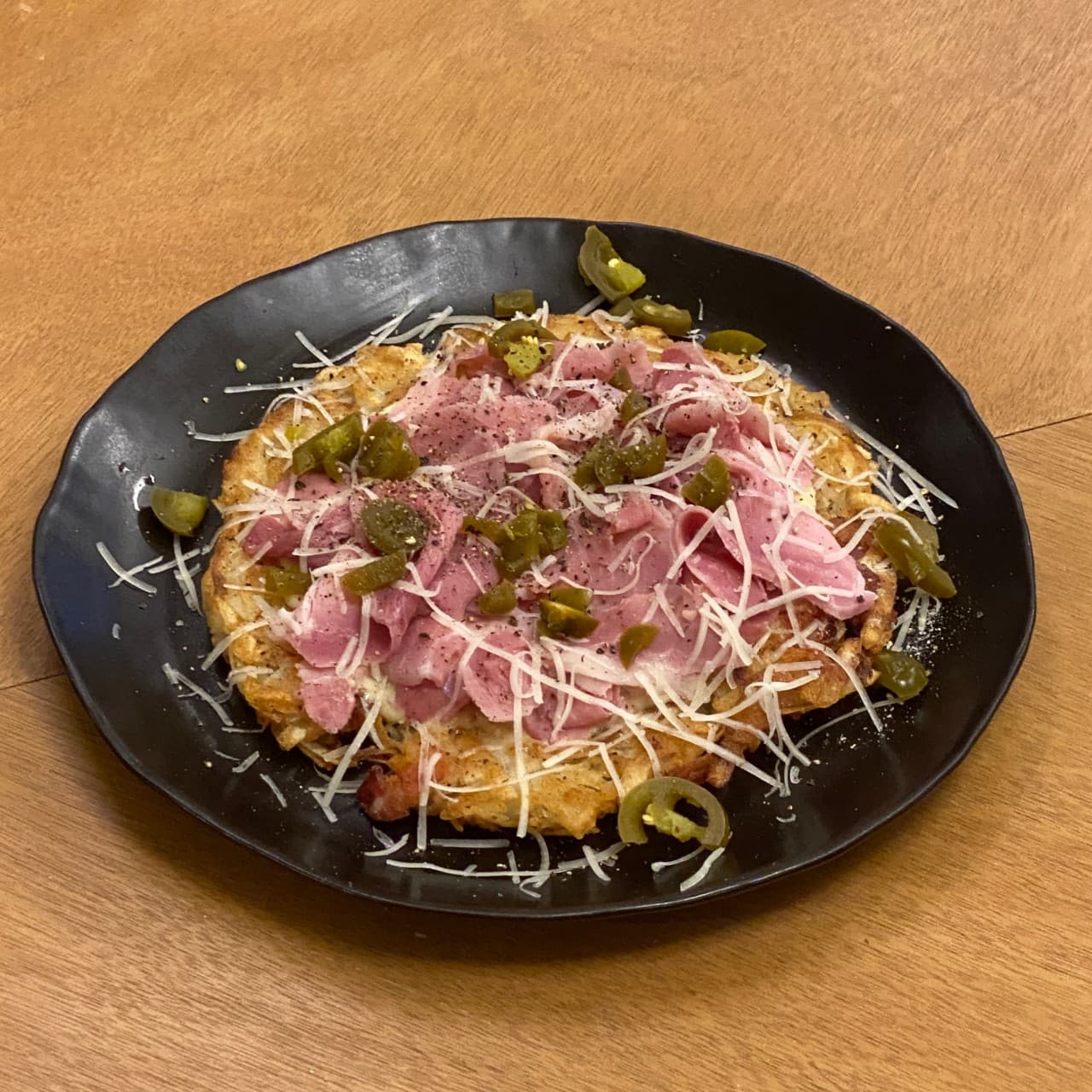 나무 소재의 바 테이블 위 검은 접시에 담긴 햄과 할라피뇨, 치즈 가루를 곁들인 감자채전 안주