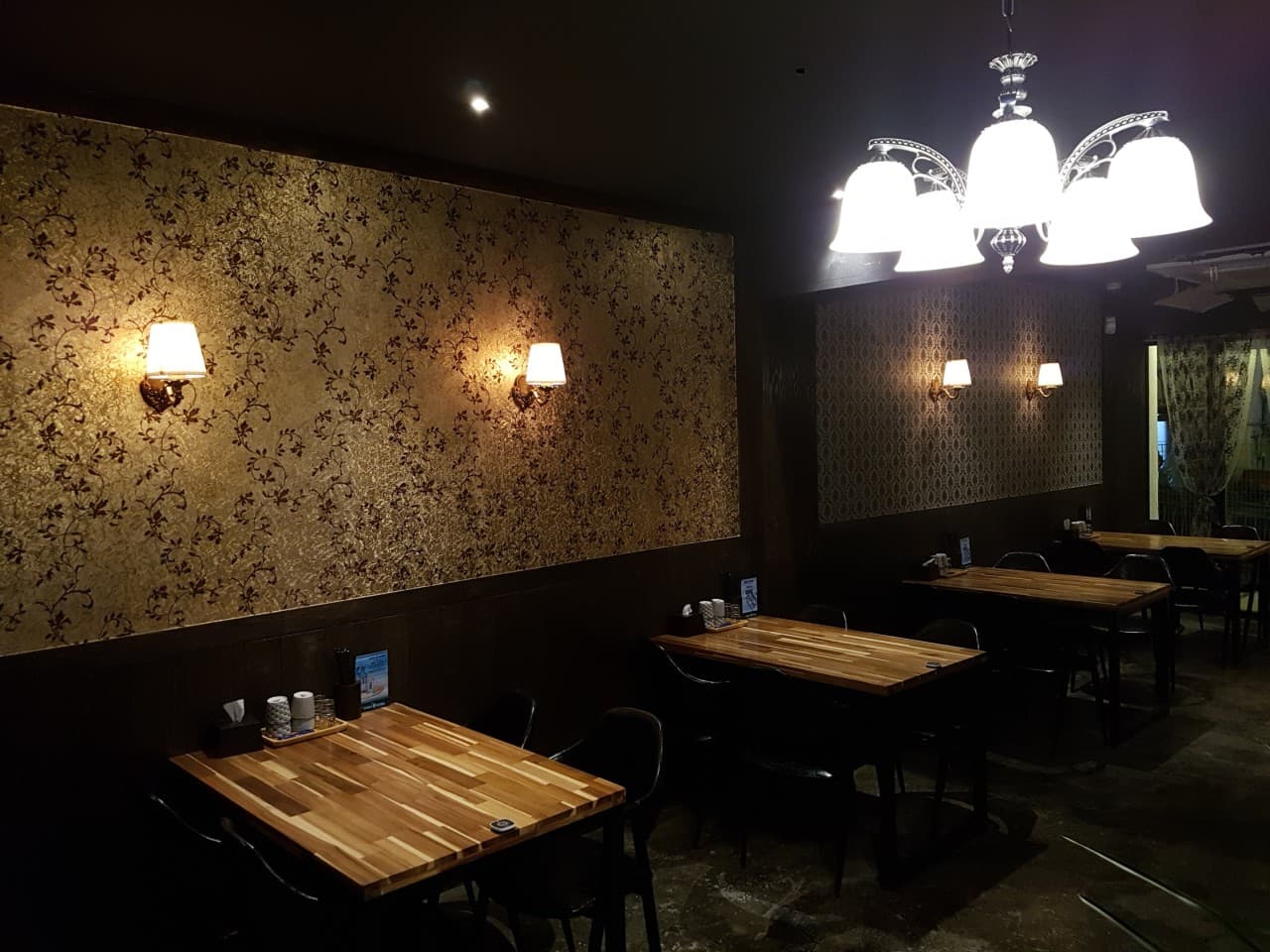 노란 조명이 비추는 패턴 벽지 아래 개별 목재 테이블과 검은 의자가 배치된 어두운 실내 전경