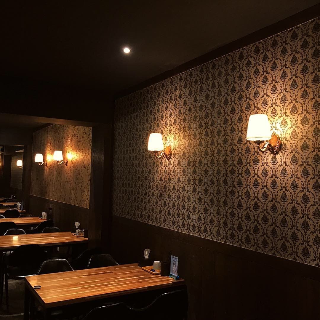 패턴 벽지와 노란 벽등 아래 배치된 소형 목재 테이블들. 거울에 반사된 어두운 조도의 실내와 1인 또는 소규모 방문객을 위한 좌석 구조.