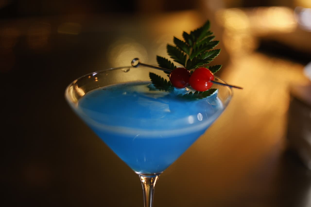 어두운 바 테이블 위 붉은 체리와 초록 잎으로 장식된 푸른색 칵테일이 담긴 마티니 잔