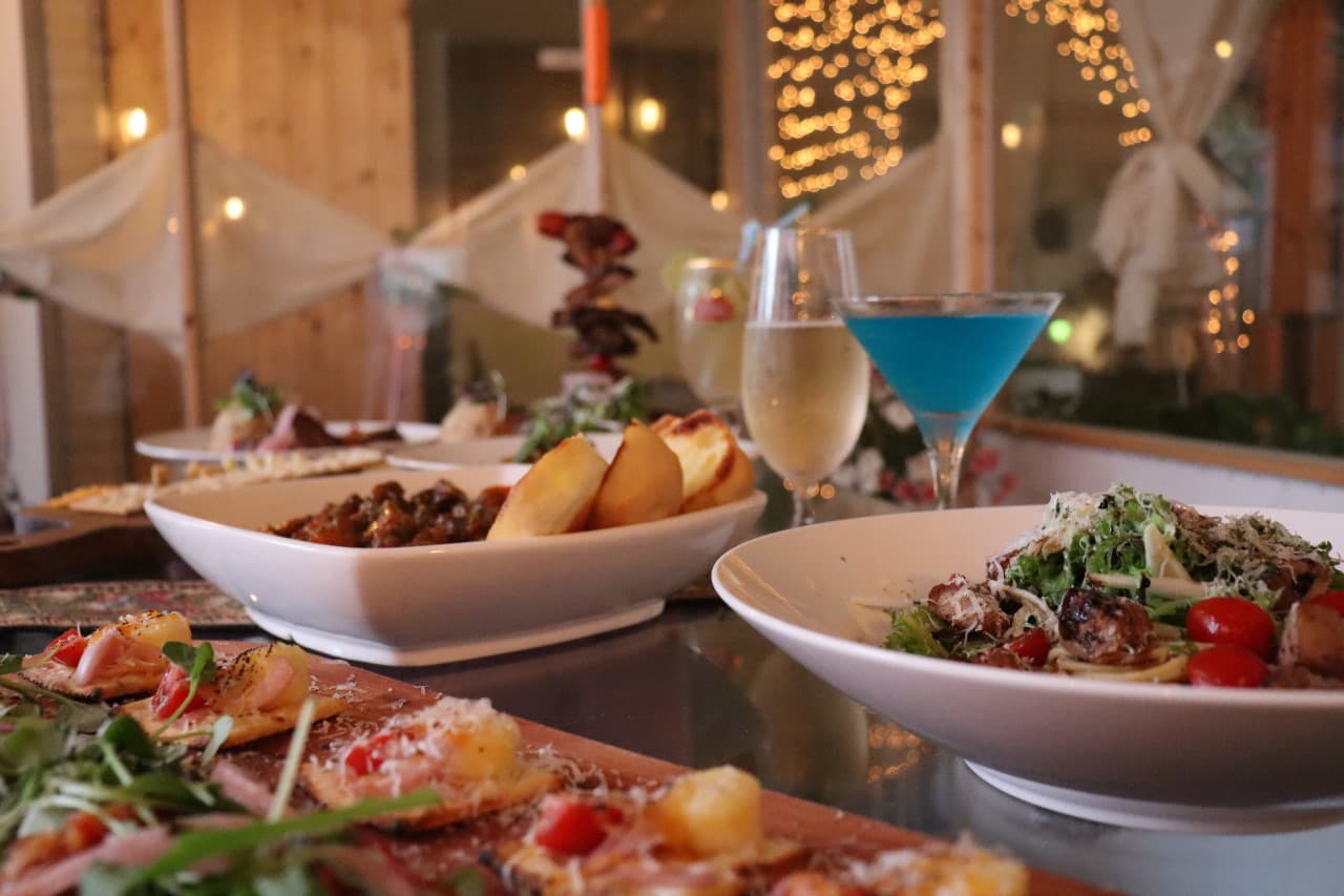 목재 테이블 위 샐러드와 카나페, 파란색 칵테일과 와인 잔이 차례로 놓인 전경. 배경에는 노란색 전구 조명이 빛나는 공간.