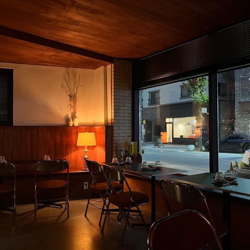 주황색 조명이 켜진 실내 창가를 따라 놓인 소형 테이블과 접이식 의자, 유리창 너머로 보이는 밤거리 풍경