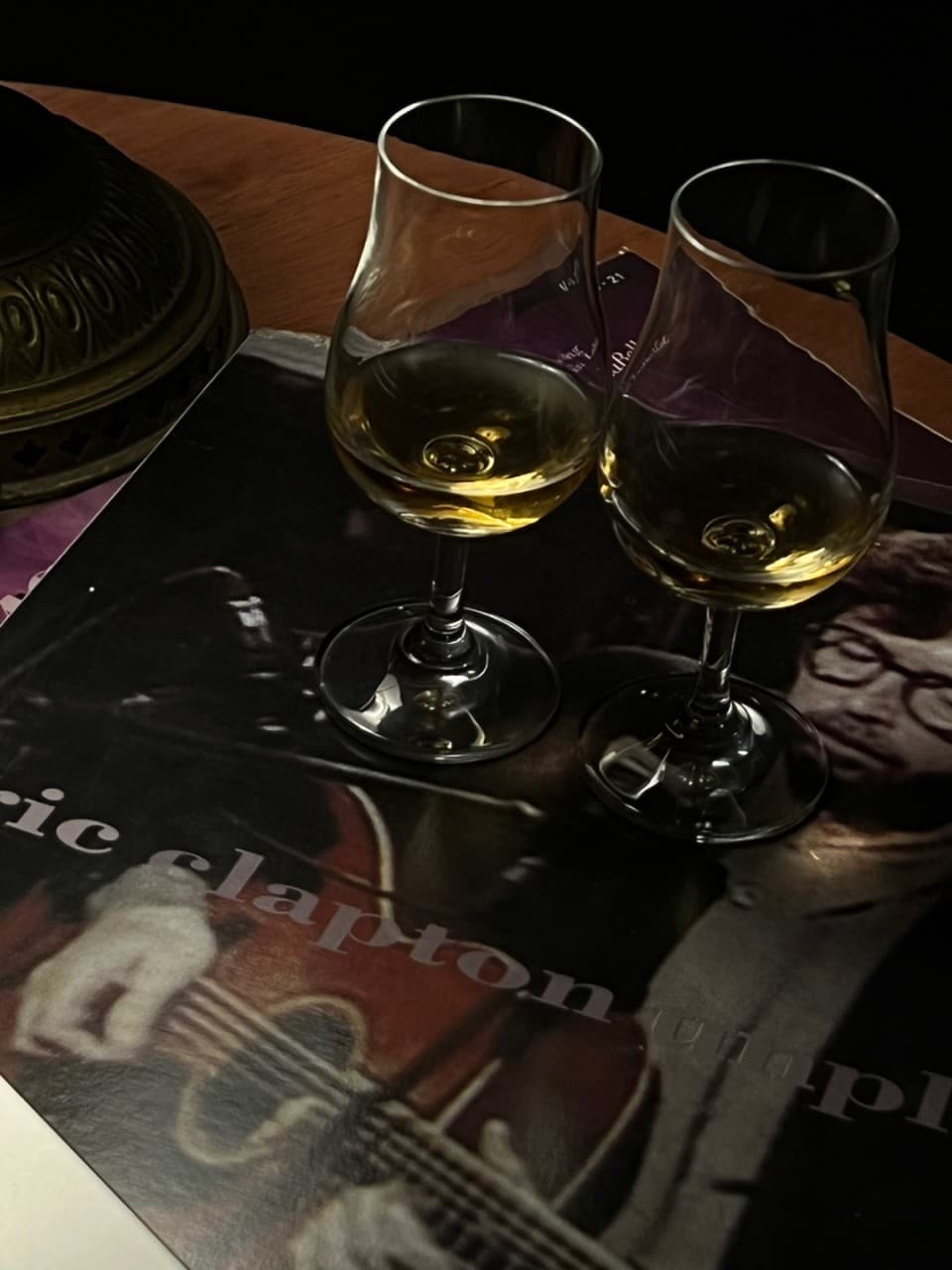 어두운 조명 아래 에릭 클랩튼 LP 커버와 그 위에 나란히 놓인 두 잔의 위스키가 보이는 목재 테이블