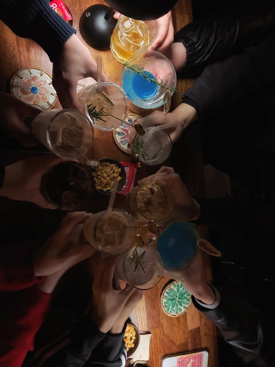 어두운 조명 아래 목재 테이블 위에서 푸른색 칵테일과 하이볼 잔을 들고 모여 있는 손들과 옥수수 안주