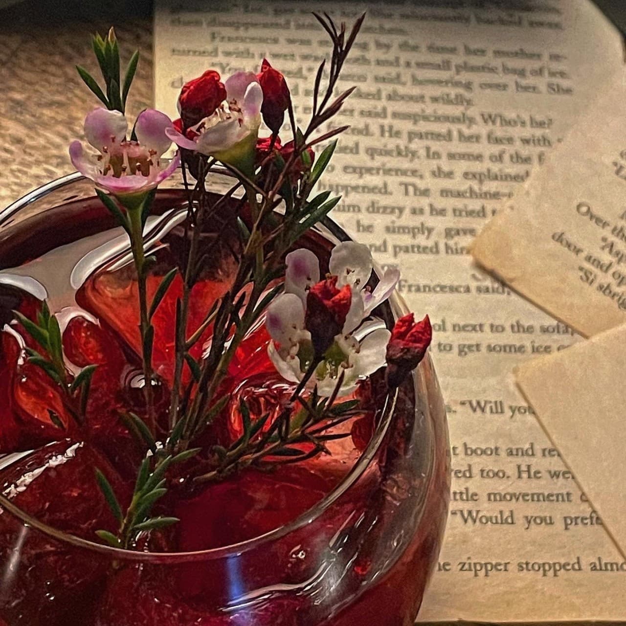 붉은색 술과 얼음이 담긴 유리잔에 꽂힌 작은 꽃 장식과 배경에 놓인 영문 서적 페이지