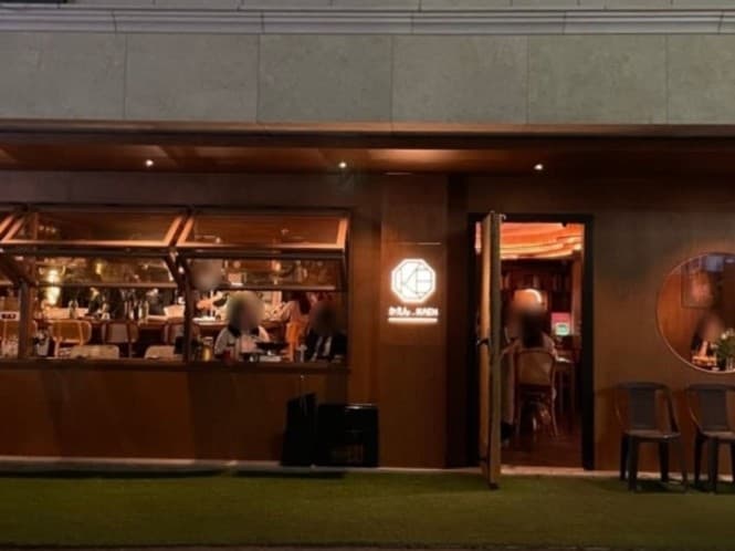 주황색 조명이 비치는 목재 외관과 개방형 창문 너머 바 테이블에 앉아 있는 사람들의 모습