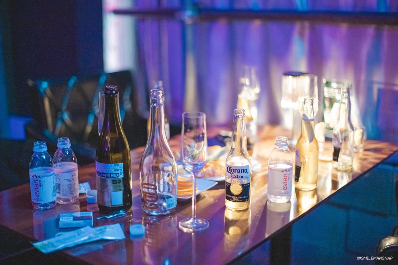 보라색 조명과 노란 불빛이 비치는 테이블 위 맥주병과 생수병, 긴 유리잔이 놓인 바 테이블 전경
