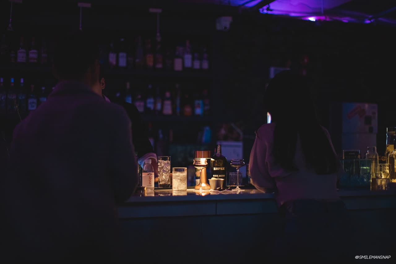 어두운 보라색 조명 아래 바 카운터에 띄엄띄엄 앉은 사람들의 뒷모습과 유리잔, 여러 종류의 술병이 진열된 선반